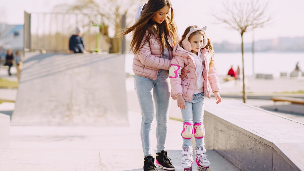 Full body focused young woman supporting daughter wearing protection pads while roller skating on concrete ramp during active leisure in sunny spring day